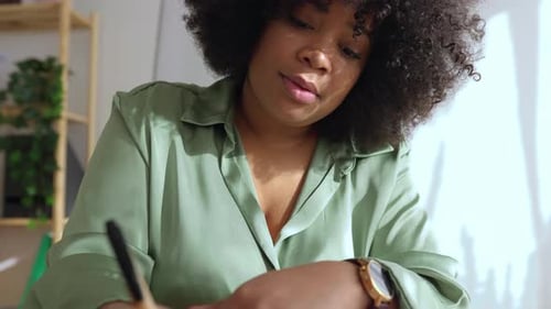 Young Black Woman Sitting at Desk Working on Laptop Writing in Paper Notebook