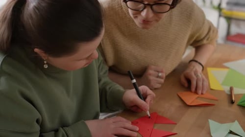 Girl Drawing on Origami Paper at Table with Woman