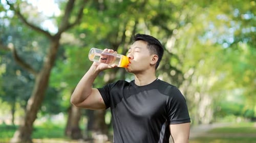 Young adult Asian athletic man drinking water from a bottle after a morning run while standing