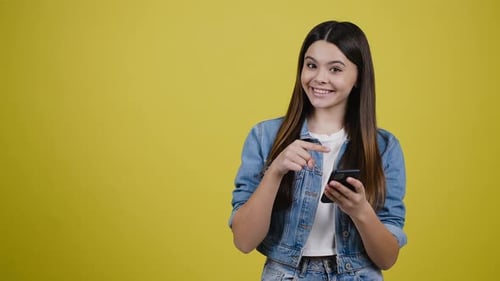 Teen Girl with Smartphone Smiles at Camera Pointing Her Finger at Copy Space on Yellow Background