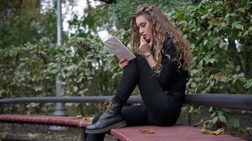 Absorbed Young Woman Reading Book Sitting on Bench in Green Park