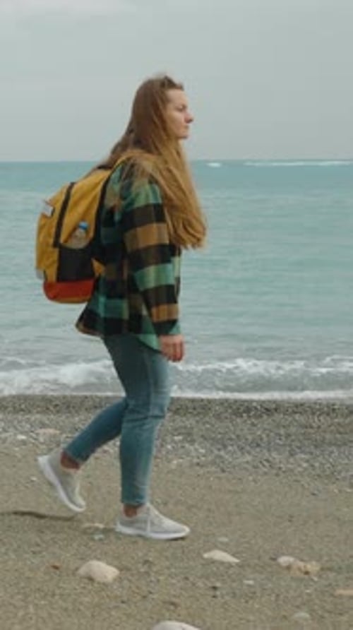 Vidéo verticale. Une jeune voyageuse avec un grand sac à dos lumineux se promène le long du bord de mer.