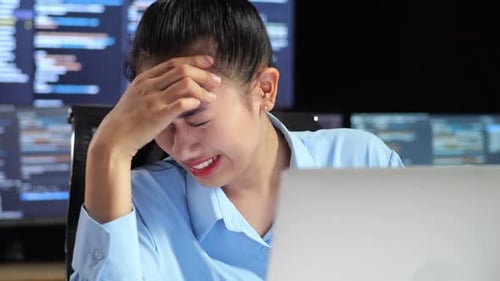 Programadora asiática que tiene dolor de cabeza mientras escribe código con una computadora portátil con varios monitores