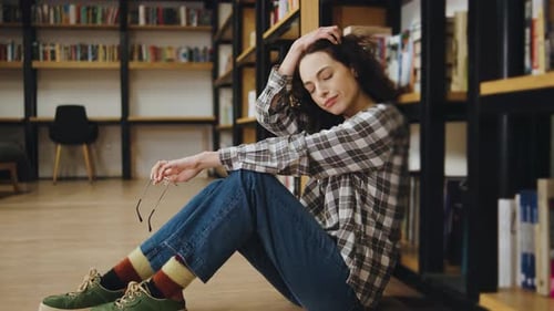 Tired Woman Sitting on the Floor of a Library Looking Stressed While Holding Glasses in a Cozy