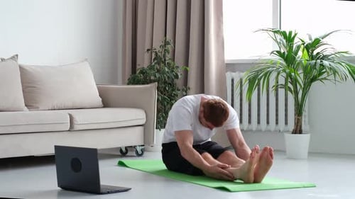 Redhead Man in White Tshirt Works Out in the Living Room Man Doing Sports on a Mat Watching Videos
