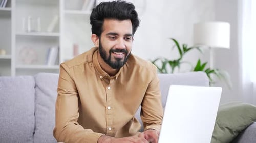Close up. Confident smiling young bearded man typing on laptop while sitting on sofa in living room