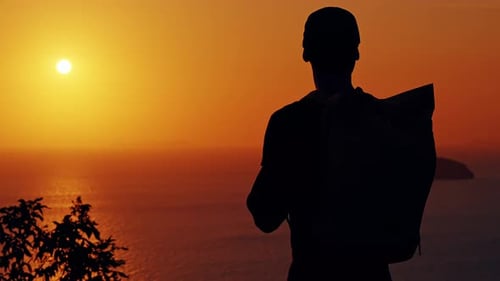 Side View Man Walking Away from Cliff Edge with Backpack During Golden Sunset with Ocean and Island