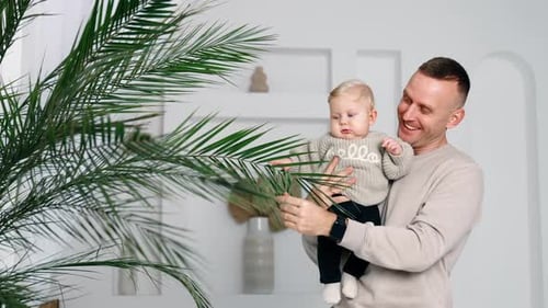 Happy dad showing his son a plant growing indoors. Baby boy looks at the tree with surprise.