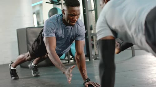 African American men performing alternating hand-tap push-ups on gym mat motivated by teamwork