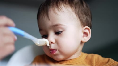 Adorable face of a lovely baby eating porridge from spoon. Feeding a kid at home. Close up.