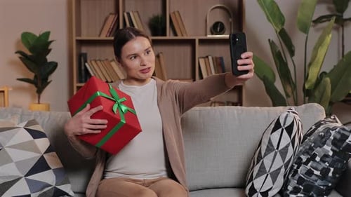 Caucasian Woman Holding Red Gift Box with Green Ribbon Taking Selfie on Smartphone Sitting on Sofa