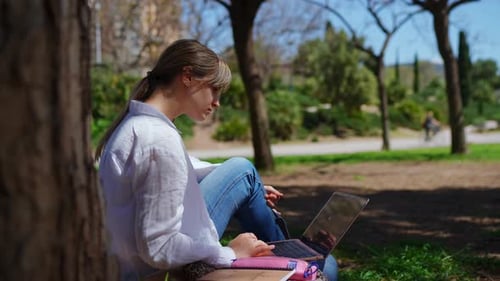 Remote Worker Sitting on Grass Typing on Laptop Amid Verdant Park Setting Sunlight Illuminating