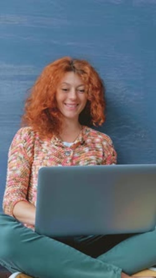 Young Woman with Curly Red Hair is Sitting on the Floor Using a Laptop Against a Textured Blue Wall