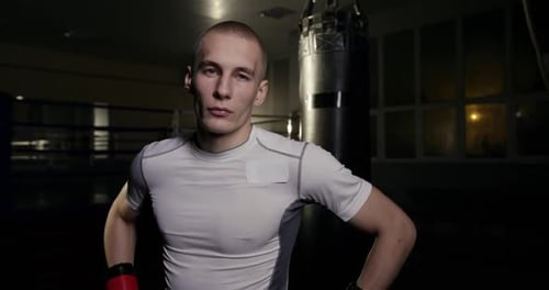 Young Guy Professional Boxer Boxing Coach Stands at Night in the Hall in Boxing Gloves Looks