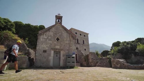 Bearded tourist with a backpack walking past an ancient stone church in Bar Fortress, Montenegro