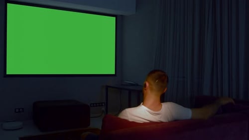 Rear view of a man sitting on sofa in living room and watching tv with green screen
