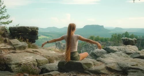 Woman with long hair practices yoga on a rocky ledge with a scenic view of the Elbe Sandstone Mounta
