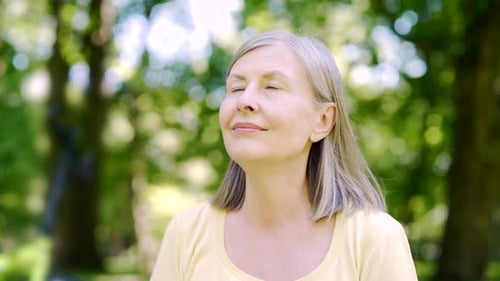 Close up portrait senior retiree woman standing in nature between forest trees relaxes, breathes