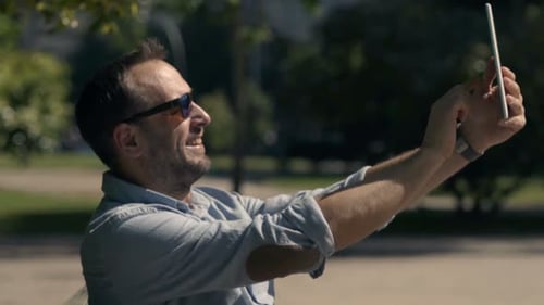 Happy Man Taking Selfie Photo with Tablet Sitting on Bench in City