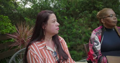 Quatre femmes assises autour d'une table en bois dans le jardin de l'arrière-cour, l'une parlant passionnément tandis que les autres sont allongées