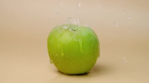 Slow motion shot of water splashing on fresh green apple on beige background.