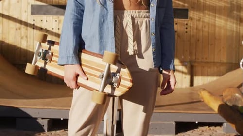 Video of midsection of caucasian female skateboarder holding skateboard in skate park
