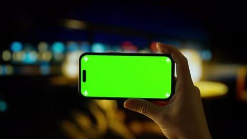Smartphone zum Halten in der Hand mit leuchtend grünem Chroma-Key-Bildschirm gegen verschwommene Nachtlichter in der Stadt