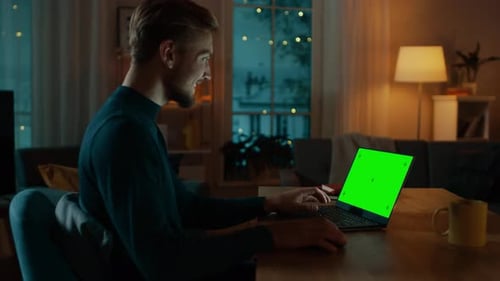 Man Sitting at His Desk Works on a Laptop with Green Mock-up Screen. Late at Night in His Living Ro