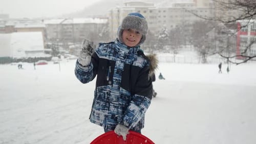Slow motion portrait of a happy smiling boy standing on a snowy hill with his plastic sleds