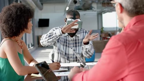 African american businessman wearing vr headset, gesturing while colleagues watching him at office