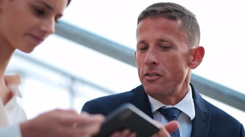 A mature businessman sits outdoors and explains something to his younger female colleague