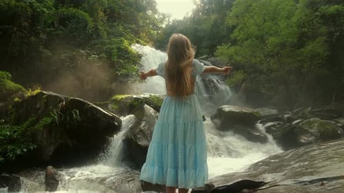 Woman in Blue Dress Stands with Arms Wide Open Near Serene Waterfall
