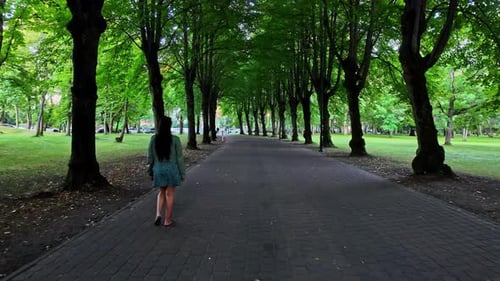 Young woman rear view walking slow motion down shaded tree footpath in public park open space