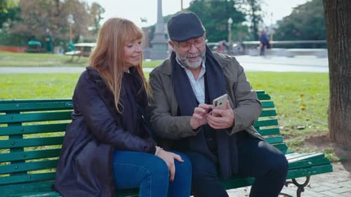 Senior Couple Using Smartphone and Talking on Bench in the Park