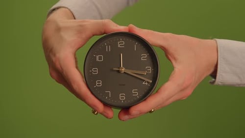 Hands Delicately Holding a Clock Against a Vibrant Green Background to Indicate Time