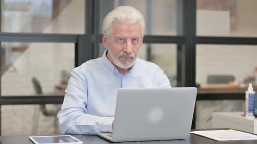 Old Businessman Smiling at Camera while Working on Laptop in Office