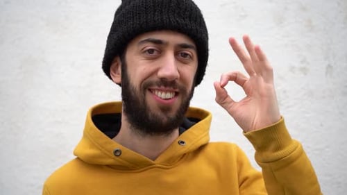 Man In Yellow Hoodie And Beanie Hat Doing An Okay Sign While Smiling In Front Of The Camera. Close-u
