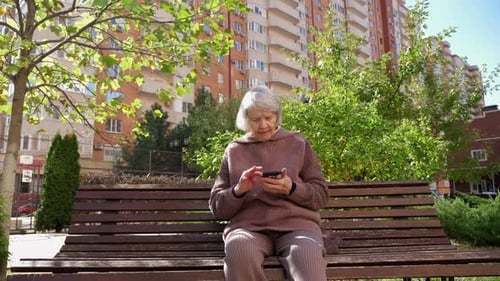An Elderly Grandmother with Gray Hair with a Phone is Sitting on a Park Bench
