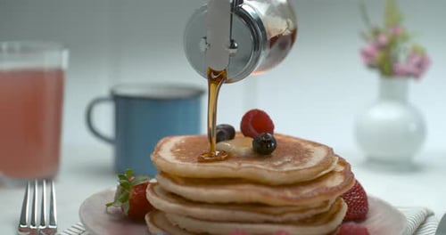 Maple Syrup Is Poured Over A Stack Of Pancakes With Fruit And Butter On A Table With A Background...