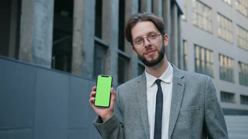 Young Caucasian Businessman in Glasses Standing Near Office Showing His Phone Phone with Green