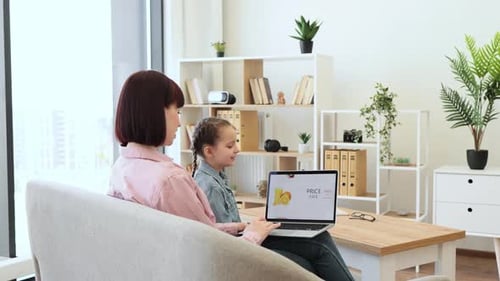 Side View of Charming Brunette Sitting on Couch with Daughter and Using Wireless Laptop