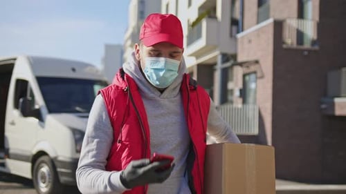 Delivery Man in Face Mask and Gloves Handling Parcel Box Walking Along the Street Talking on Phone