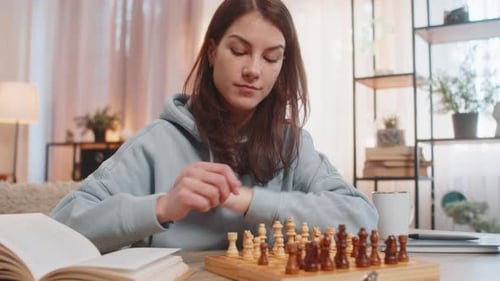 Young Woman Playing Chess Alone Arranging Pieces at Table on Sofa with Book Lying Nearby at Home