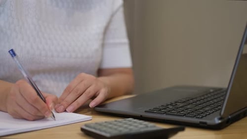 Woman Writes on Notepad with Laptop and Calculator