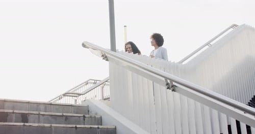 Diverse female friends pausing to gaze skyline from overpass, holding coffee, descending stairs