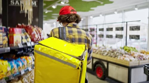 Rear View of a Confident Brunette Guy with Curly Hair in a Checkered Shirt with a Yellow Bag a Food
