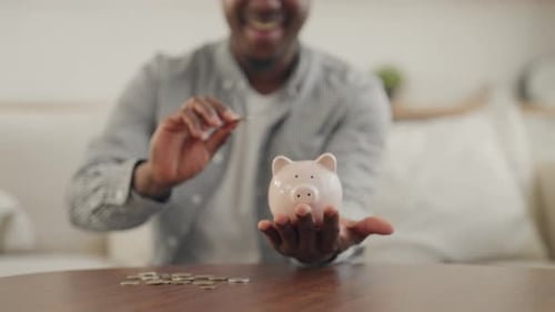 Young Adult Black Man Putting Coins in Piggy Bank Closeup of Hands at Home Saving Money and Future