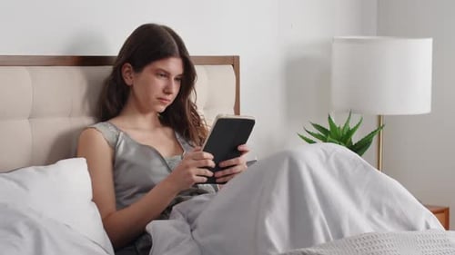 View of a Young Pleasant Woman Who Lies in Bed and Looks at Something on a Digital Tablet A Brunette