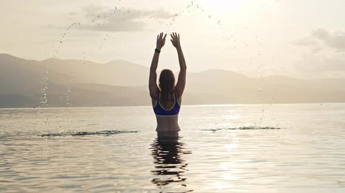 Happy Young Woman Stands in Ocean and Raises Her Hands Up with Splashes From Water at Sunset