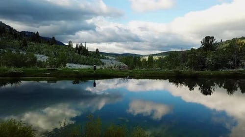 A drone shot backing up from a pond with a green meadow and mountains seen surrounding it. (slow mot